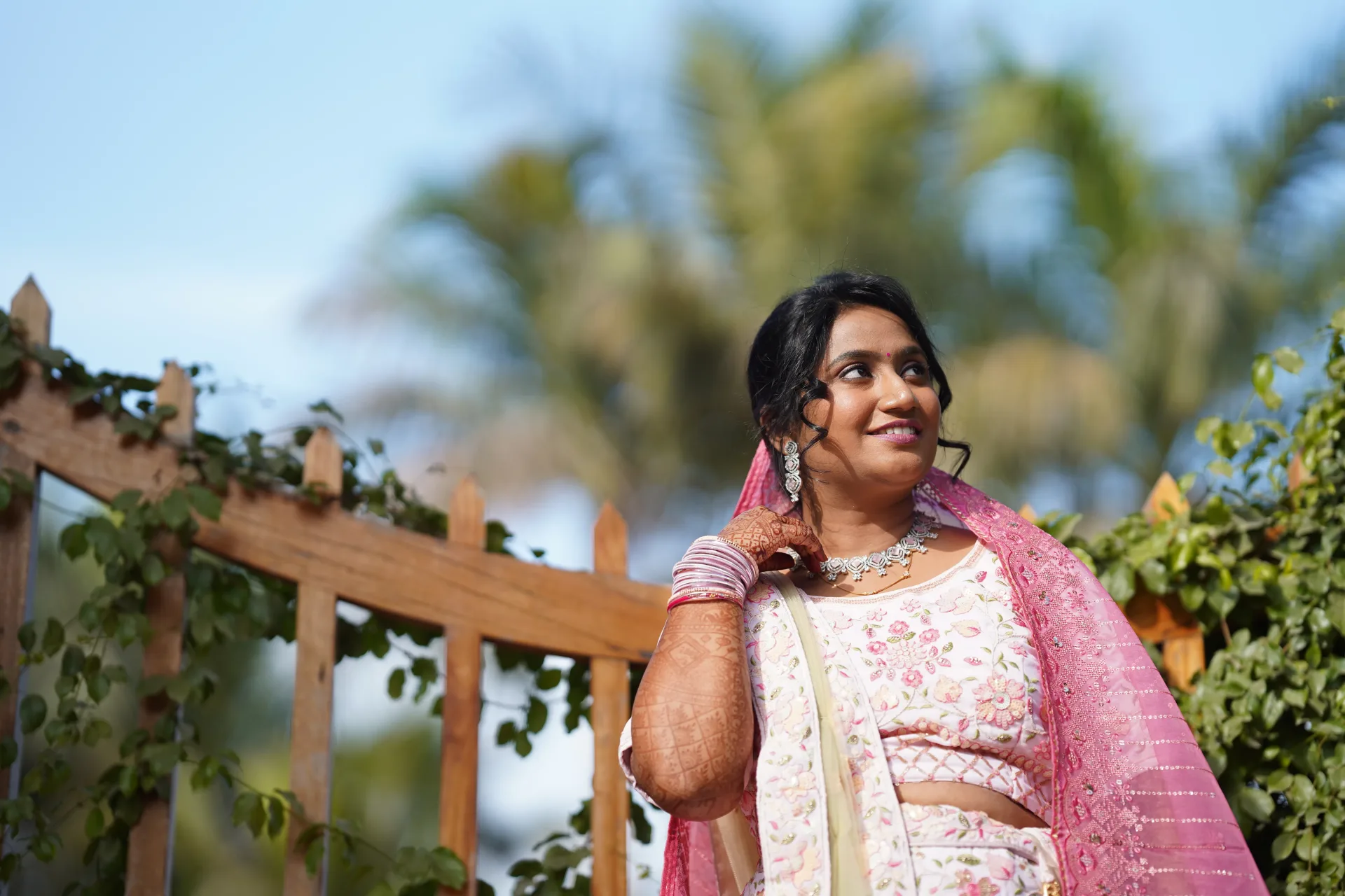 A bride in traditional attire poses gracefully near a wooden fence.
