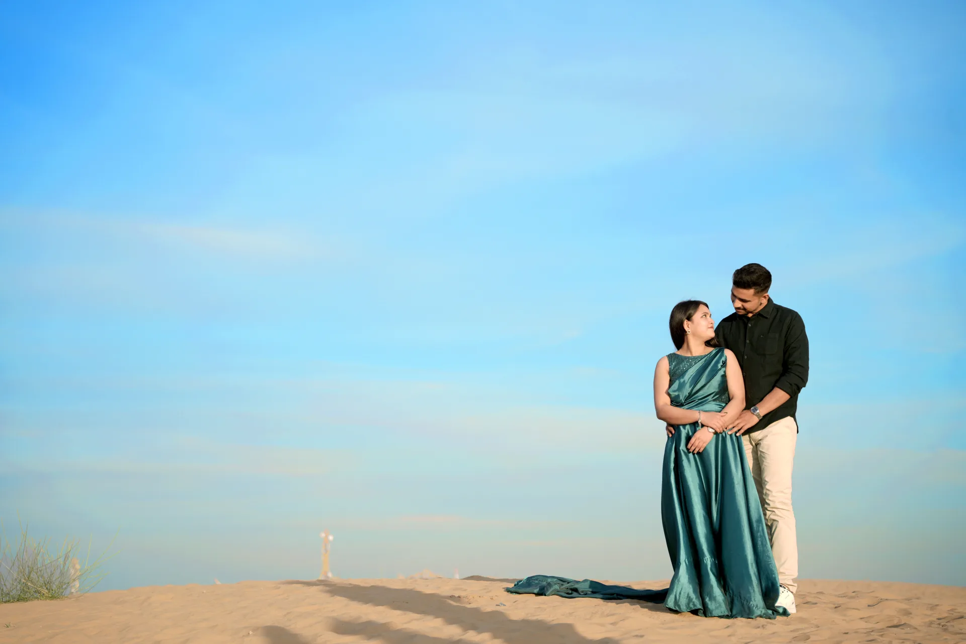 Couple posing in a sandy area under a clear blue sky for a pre-wedding shoot.