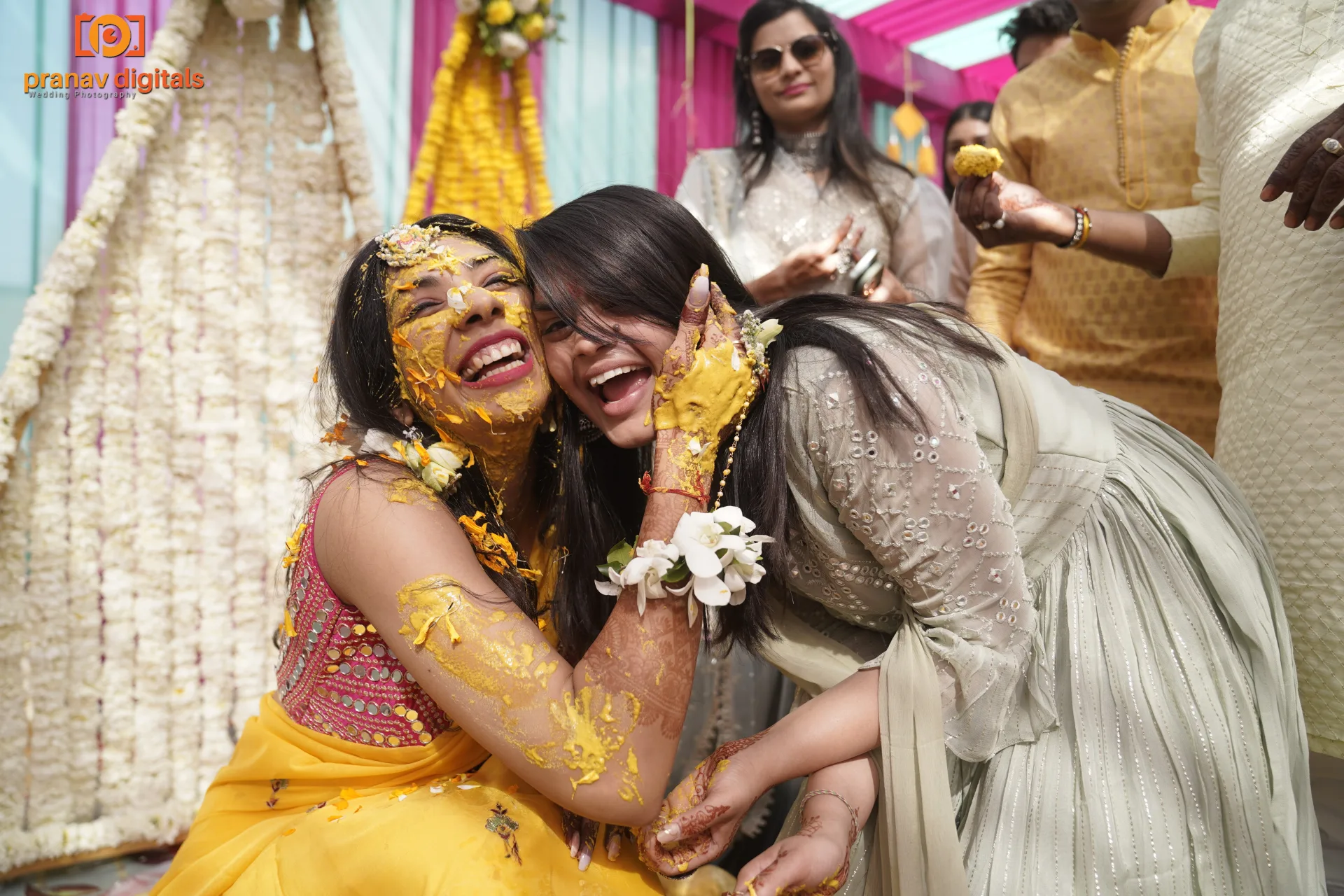 Two women laughing and celebrating during a festive ceremony covered in colorful powders.